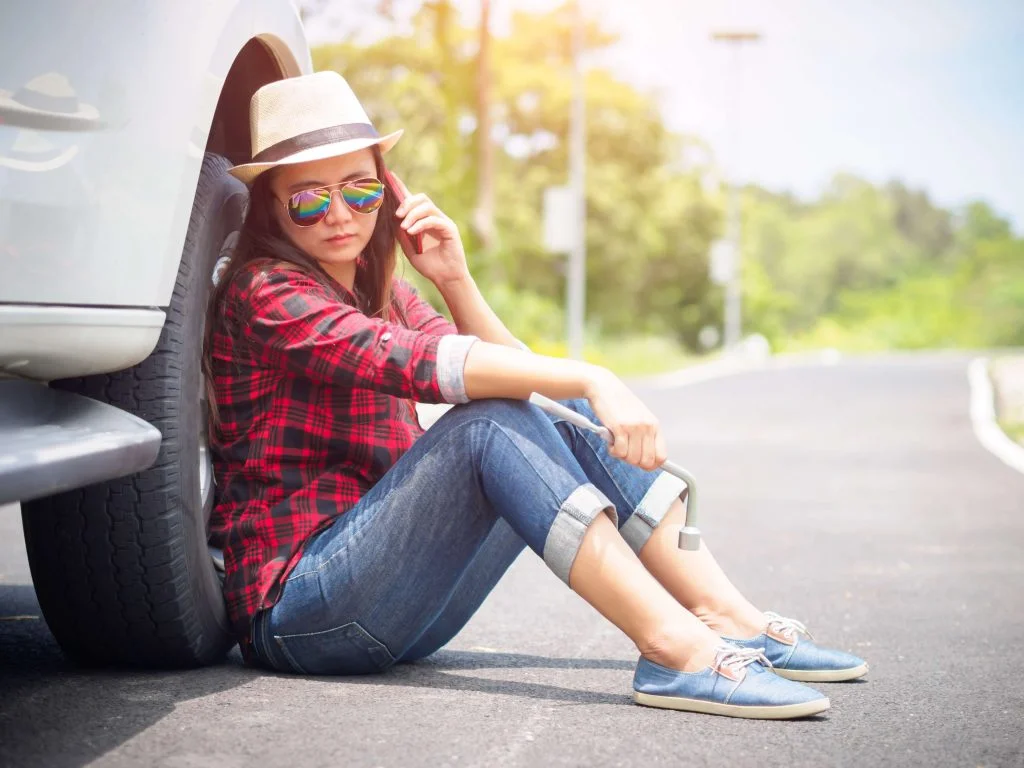 photo of a woman on the phone stuck on the side of the road with a wrench in her hand